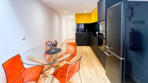 A modern kitchen with a black refrigerator, a glass dining table with orange chairs, and a yellow accent wall.