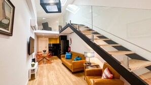 A modern apartment with a wooden floor, a staircase with glass railings, a living room with a sofa and a coffee table, and a kitchen in the background.