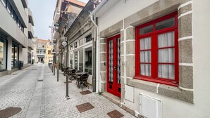 A narrow cobblestone street lined with buildings, some with red-framed windows and doors, and outdoor seating areas with tables and chairs.