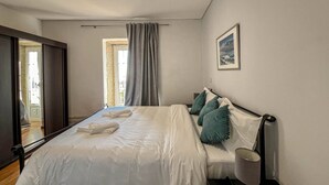 Una cama bien tendida con ropa blanca y almohadas color turquesa, una lámpara de noche y un cuadro en la pared.