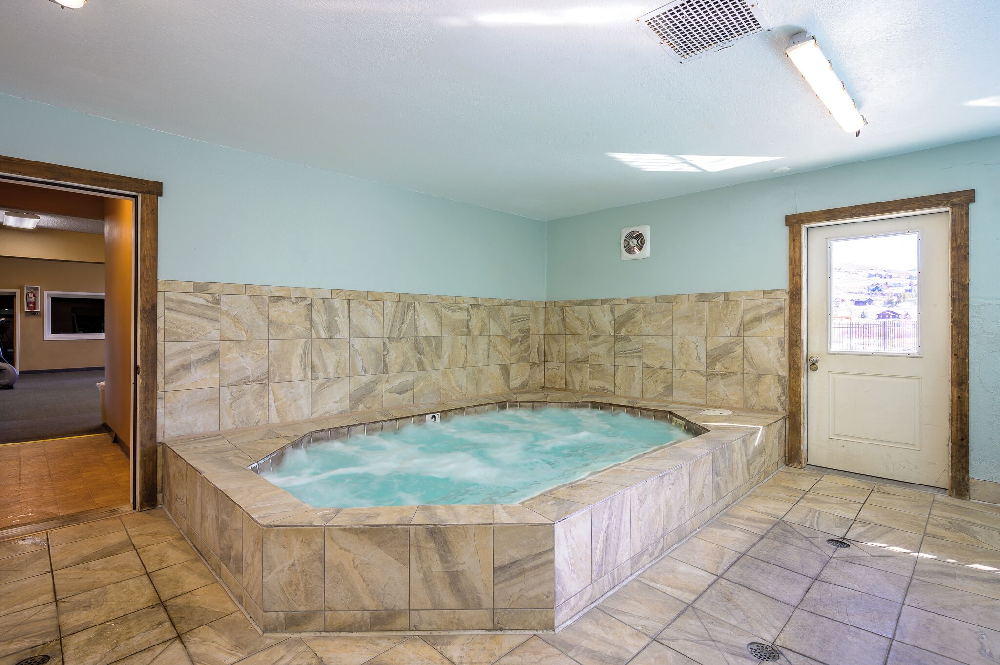 A bathroom with a marble tub, a door, and a fan on the wall.