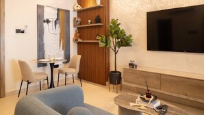 A modern living room with a grey sofa, a round coffee table, a TV mounted on the wall, a wooden panel wall, and a small dining area with a table and chairs. - Belfast studio-Blue Waters Property. (Accra)
