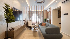 A modern living room with a grey sofa, a coffee table, and a TV mounted on the wall. There is a plant in a black pot and a wooden bench along the wall. - Belfast studio-Blue Waters Property. (Accra)