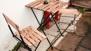 Ein Holz-Gartentisch mit Stühlen vor einer weißen Wand, im Hintergrund eine Metallbank und ein Holztor.