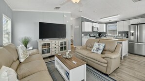 A modern living room with a flat-screen TV, beige sofas, a coffee table, and a kitchen area with stainless steel appliances. - Limited-Time Spring & Summer Deals: Dog-Friendly 2BR Beach Getaway (Jensen Beach)