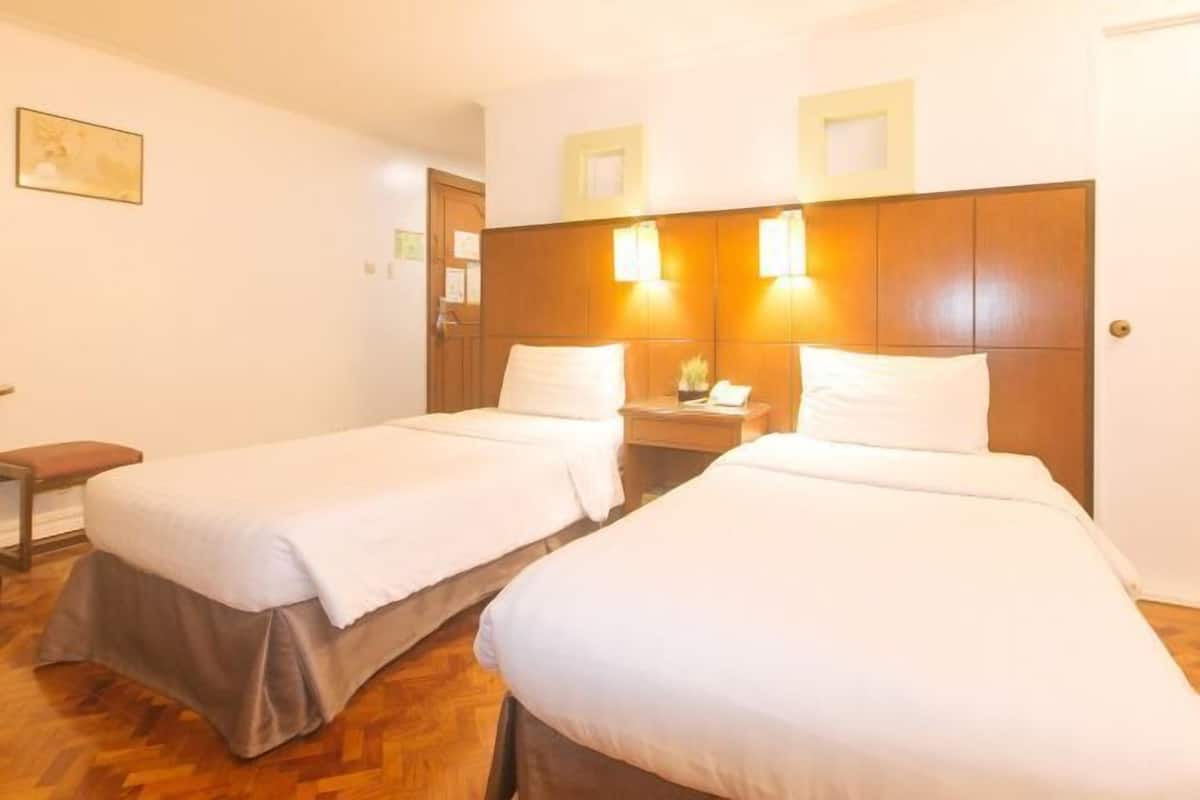 Habitació d'hotel amb dos llits individuals, panell de fusta al capçal, taules de nit i un petit banc.