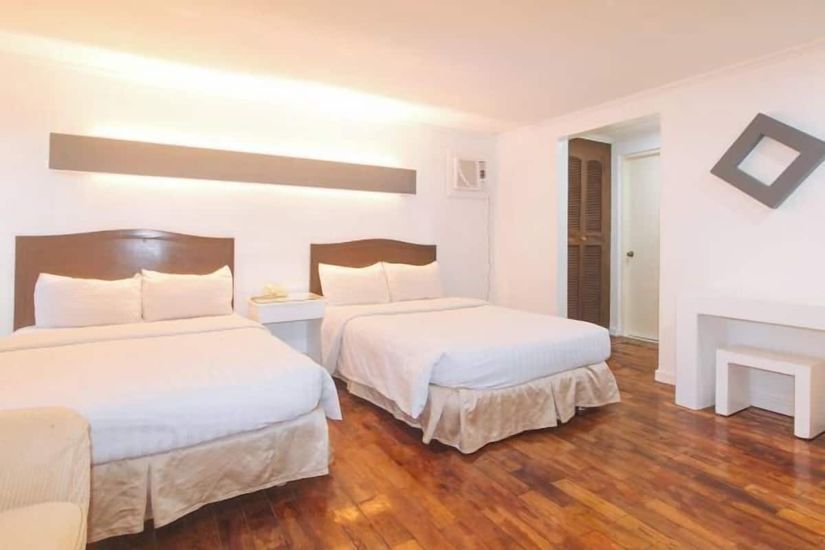 Habitació d'hotel amb dos llits, terra de fusta, una tauleta de nit amb una làmpada i un quadre al mur.