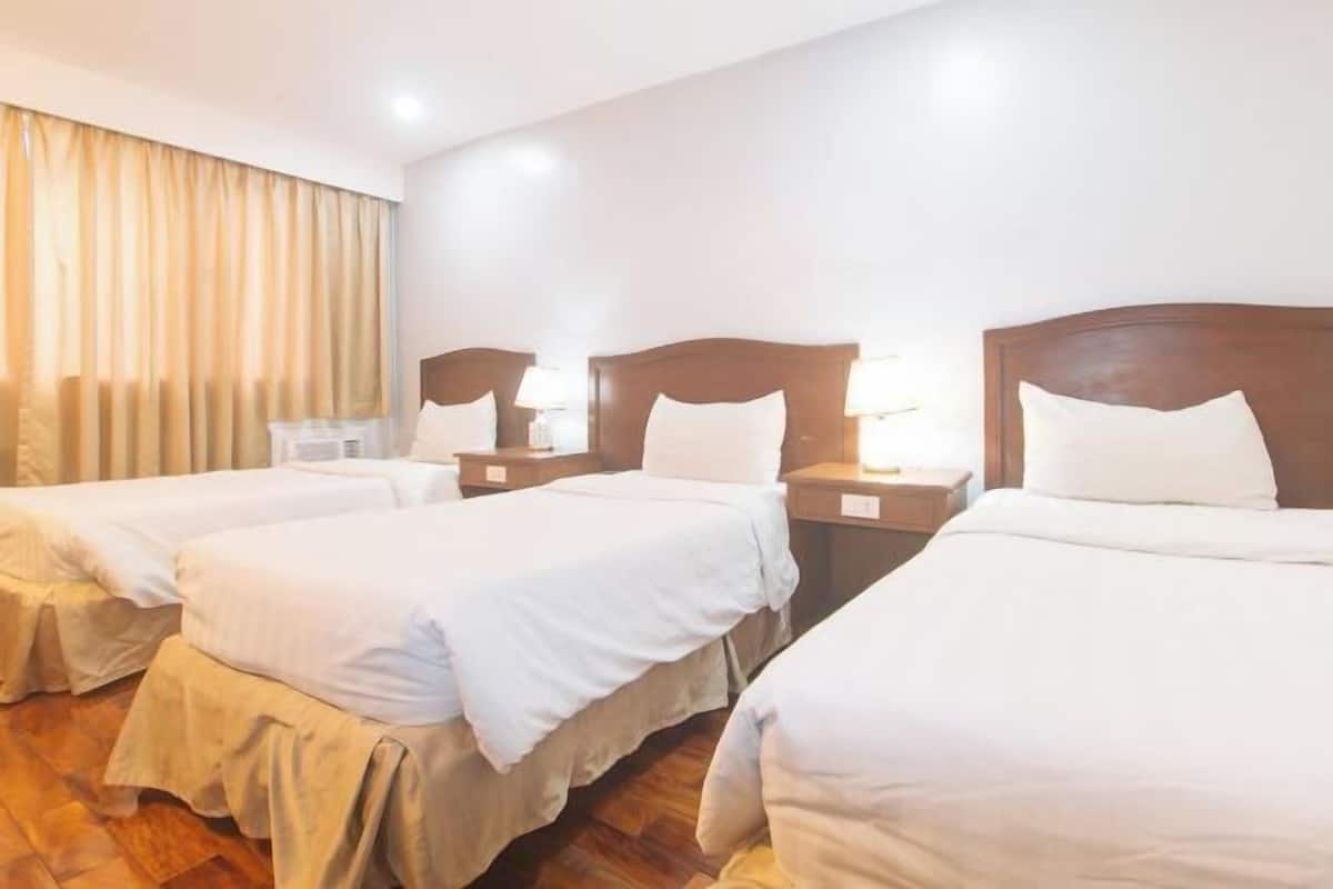 Habitació d'hotel amb tres llits individuals, amb reposabrises de fusta, taules de nit amb làmpades i un gran finestral amb cortines.