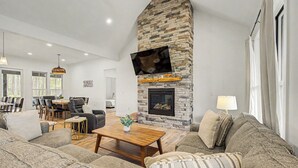 A living room with a stone fireplace, a flat-screen TV mounted above it, a sofa, a coffee table, and a dining area in the background. - Holiday House by Taylor Made Deep Creek (McHenry)