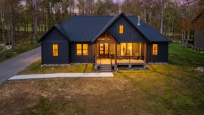 A dark-colored two-story house with a porch, a driveway, and a forested backdrop. - Holiday House by Taylor Made Deep Creek (McHenry)