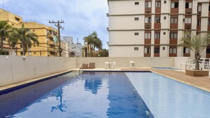 A swimming pool with a tiled deck, surrounded by buildings and palm trees. - Apto c Piscina e Brinquedoteca a 400m da Praia (Guarujá)
