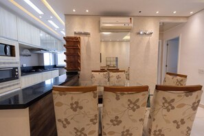 A modern kitchen with built-in appliances, a dining area with floral chairs, and an air conditioning unit. - Apto c Piscina e Brinquedoteca a 400m da Praia (Guarujá)