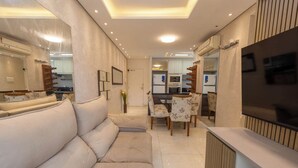 A modern living room with a sofa, dining table, chairs, and a TV. - Apto c Piscina e Brinquedoteca a 400m da Praia (Guarujá)