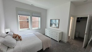 A bedroom with a bed, a dresser, a mirror, and a window with blinds. - Downtown Bridge St Apartment (Charlevoix)