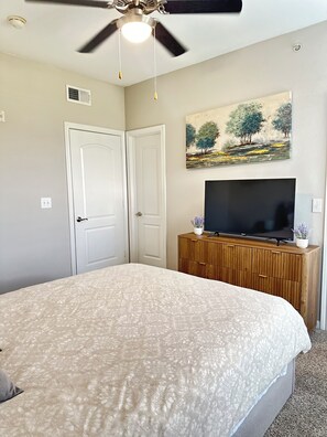 Une chambre à coucher avec un lit, un ventilateur de plafond, un téléviseur posé sur une commode en bois et un tableau accroché au mur.