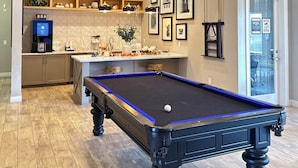A pool table in a room with a kitchen area, wooden cabinets, and a coffee machine.