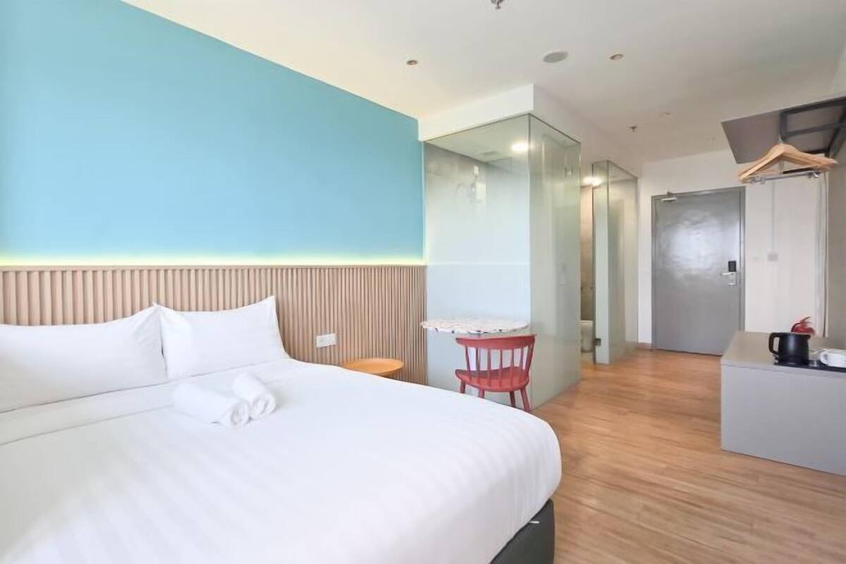 A hotel room with a bed, a small table, a chair, a mirror, and a kitchenette area.