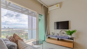 Uma sala de estar moderna com sofá, televisão fixada na parede, uma pequena mesa redonda e vista para a paisagem urbana através de amplas janelas.