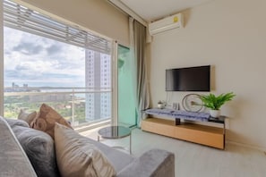 Una sala de estar moderna con un sofá, un televisor montado en la pared, una pequeña mesa redonda y vistas a un paisaje urbano a través de ventanas amplias.