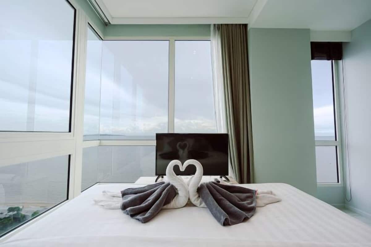 A hotel room with a bed, a heart-shaped towel arrangement, a flat-screen TV, and large windows.