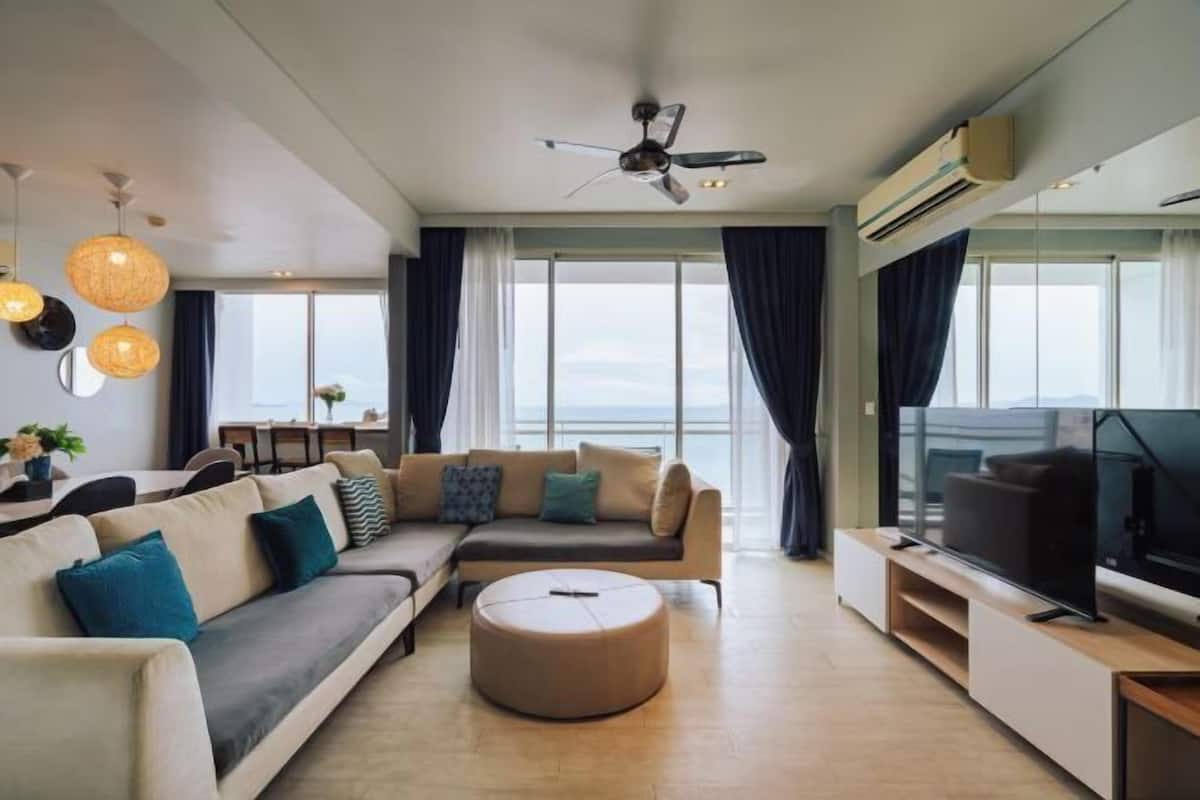 A modern living room with a sofa, ottoman, TV, ceiling fan, and large windows.