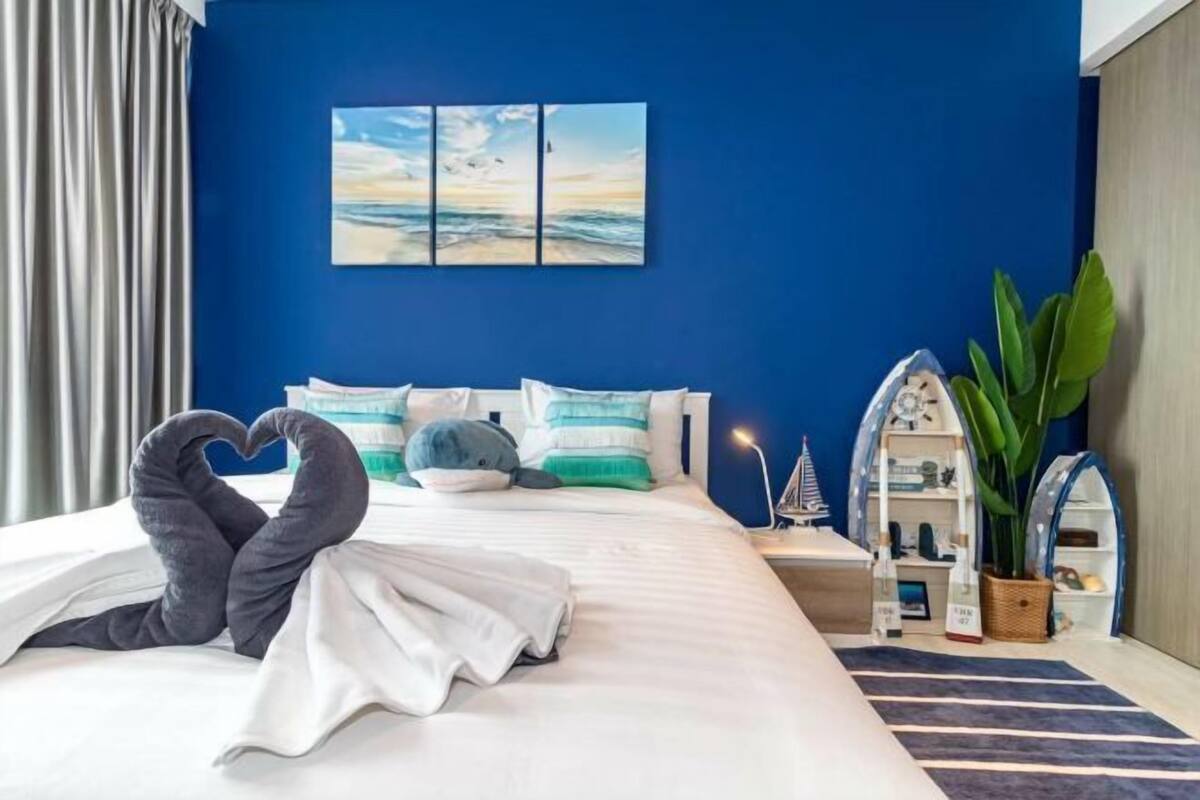 A hotel room with a large bed, a heart-shaped towel arrangement, a painting of a beach scene, a plant, and a small shelf with items.