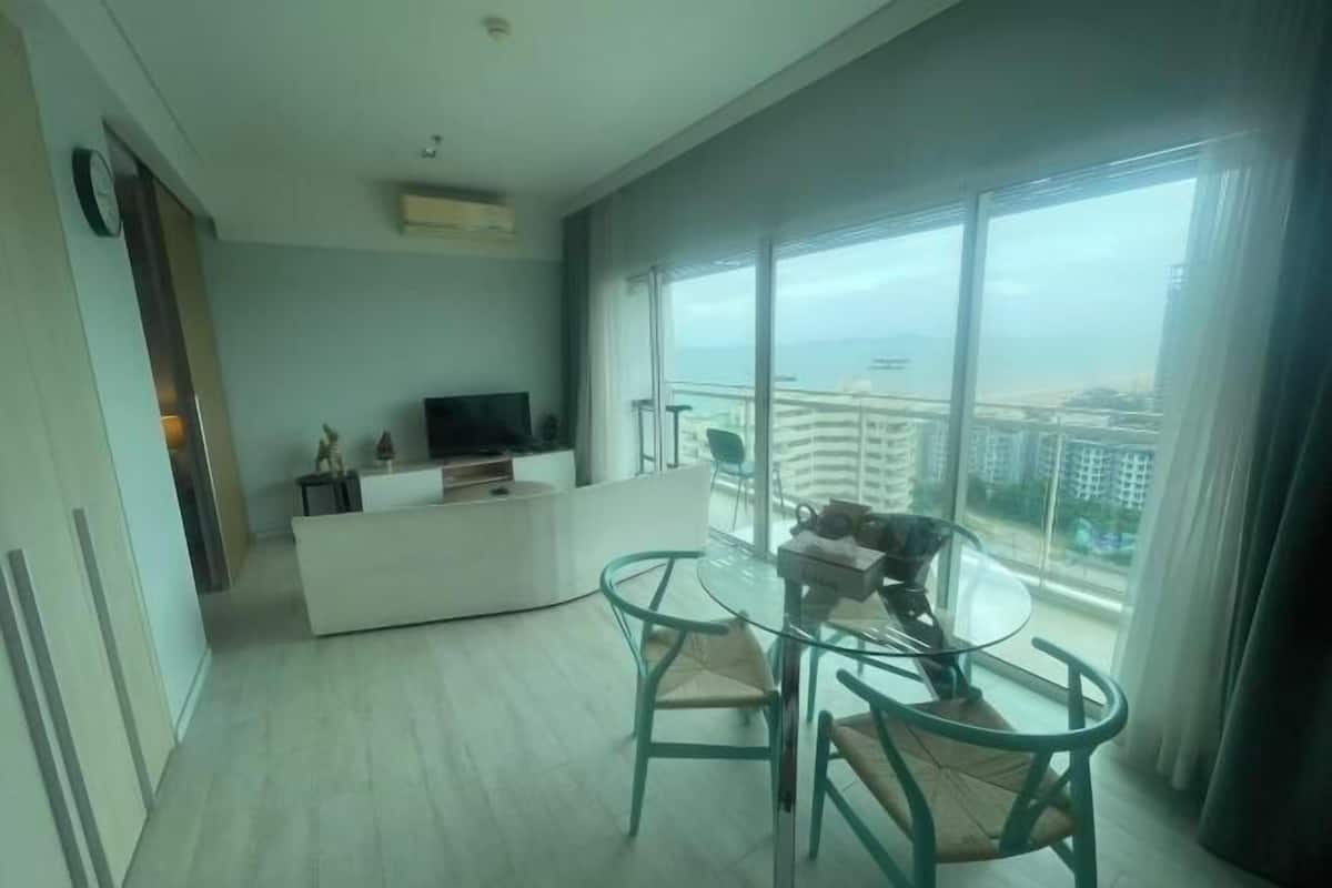 A modern living room with a glass dining table, two chairs, a white entertainment unit, and a large window with a city view.
