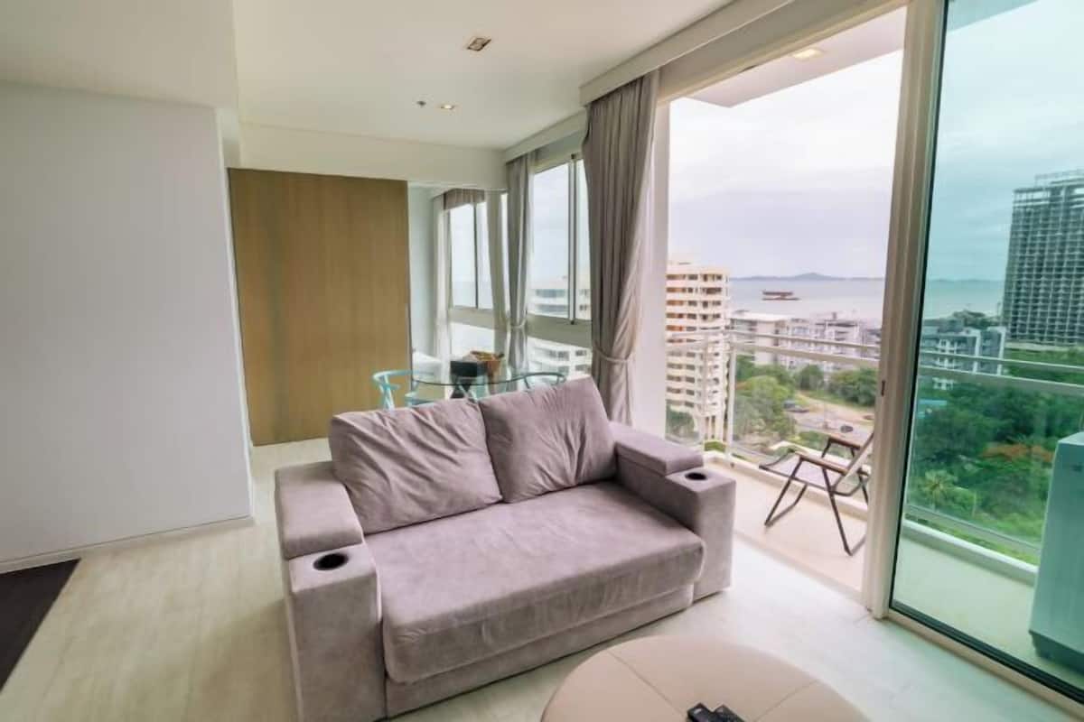 A modern living room with a grey sofa, a glass table, and a view of the city and sea through large windows.