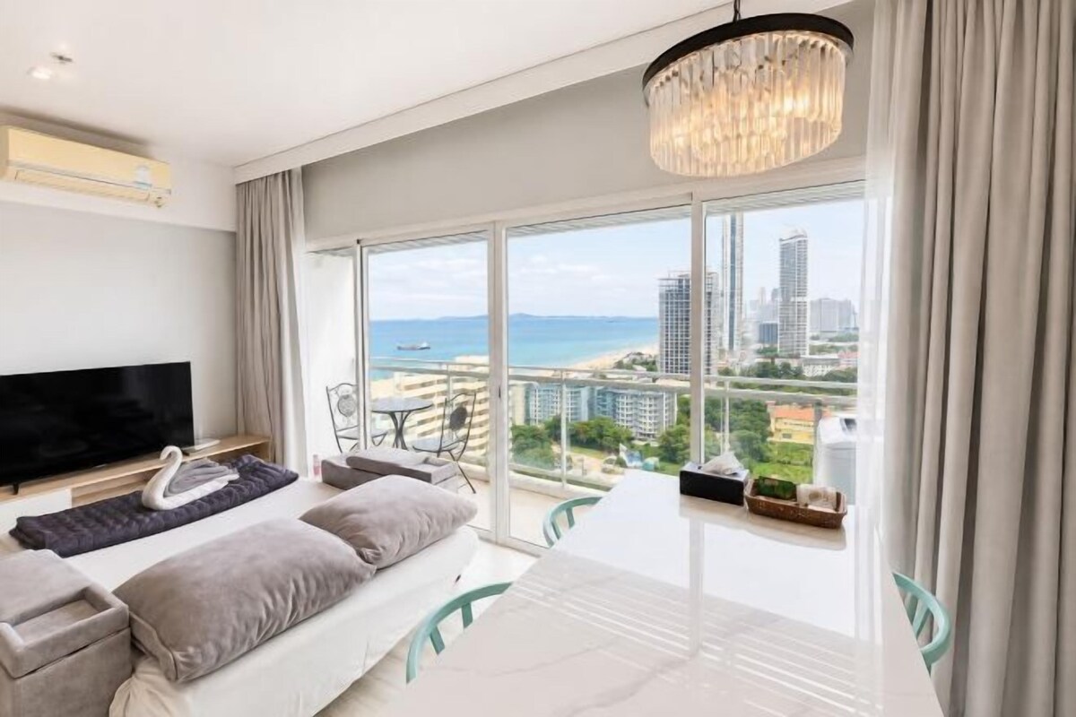 A modern hotel room with a large balcony, a flat-screen TV, a sofa, and a dining table.