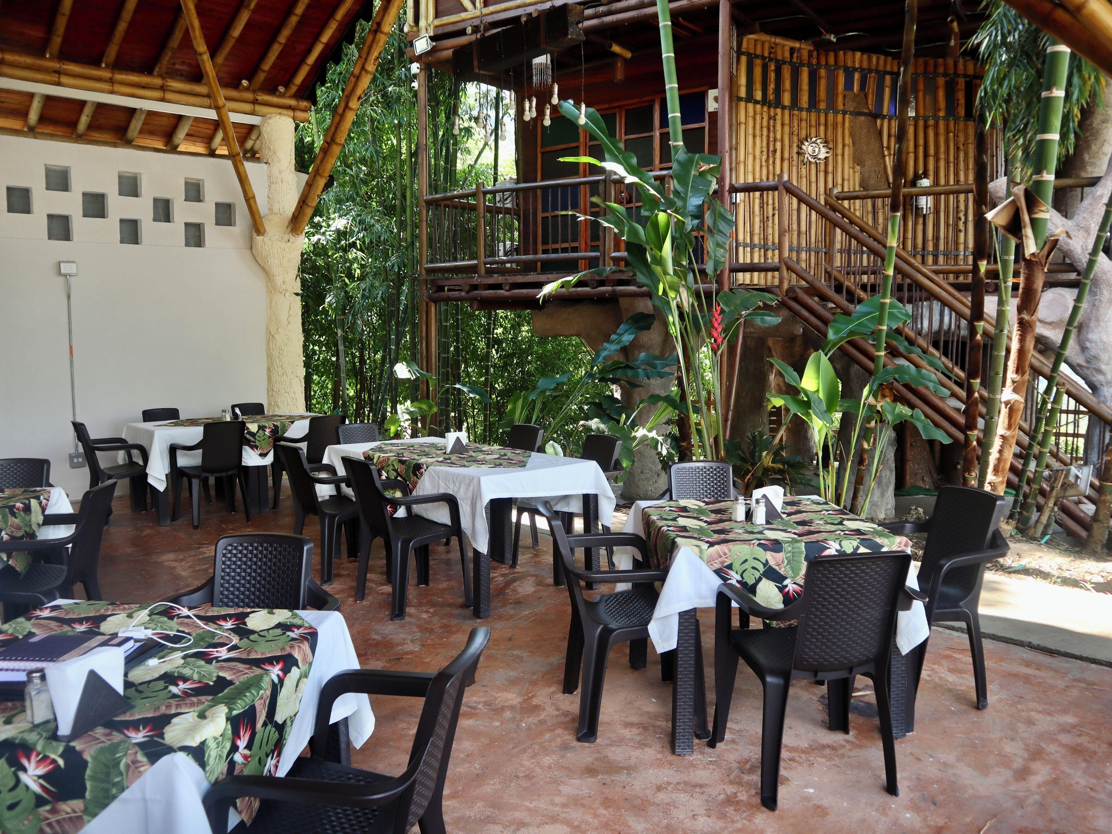 An outdoor restaurant with tables and chairs, surrounded by bamboo and tropical plants.