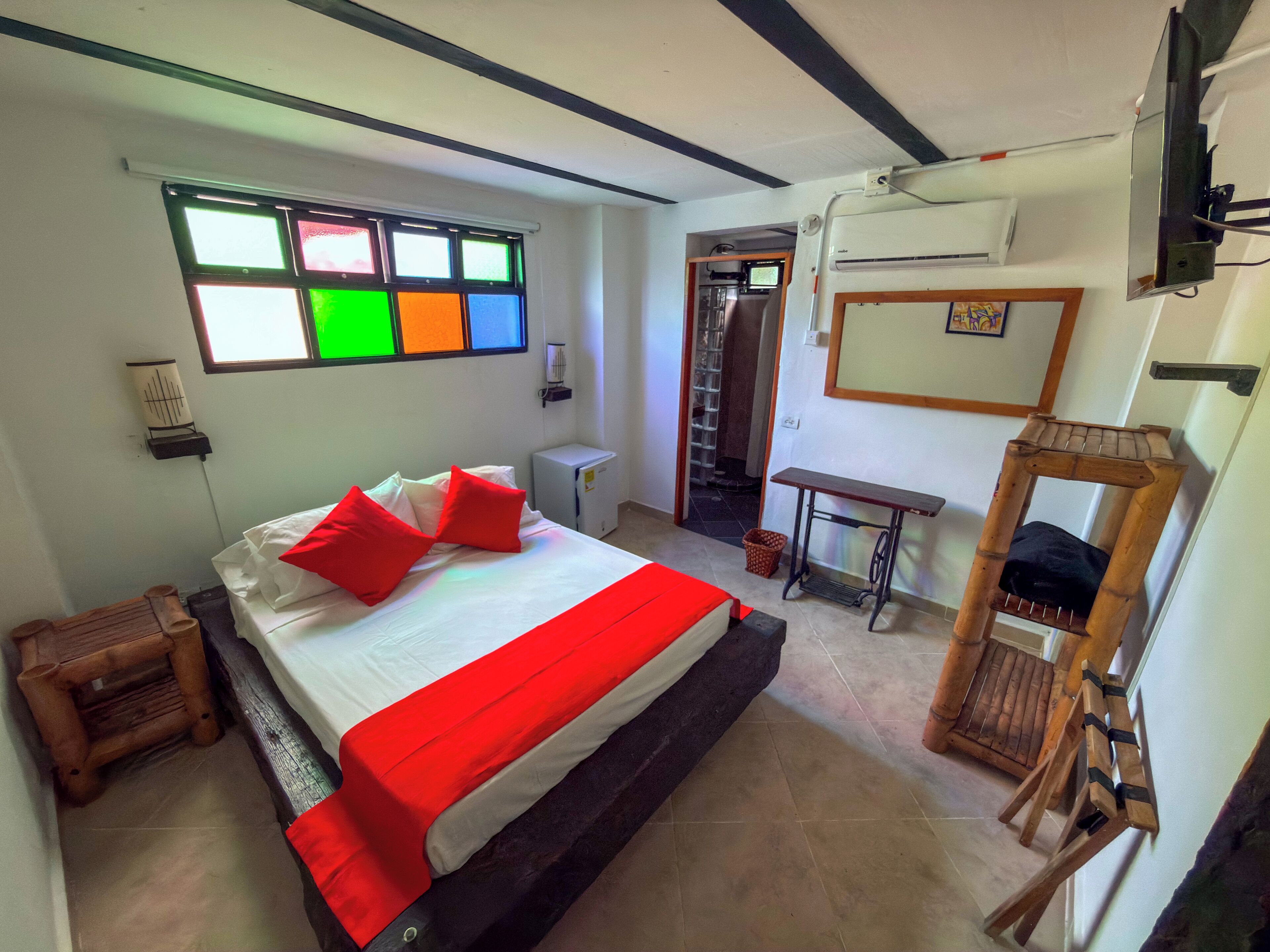 A bedroom with a bed, a small table, a mirror, and a window with colorful panes.