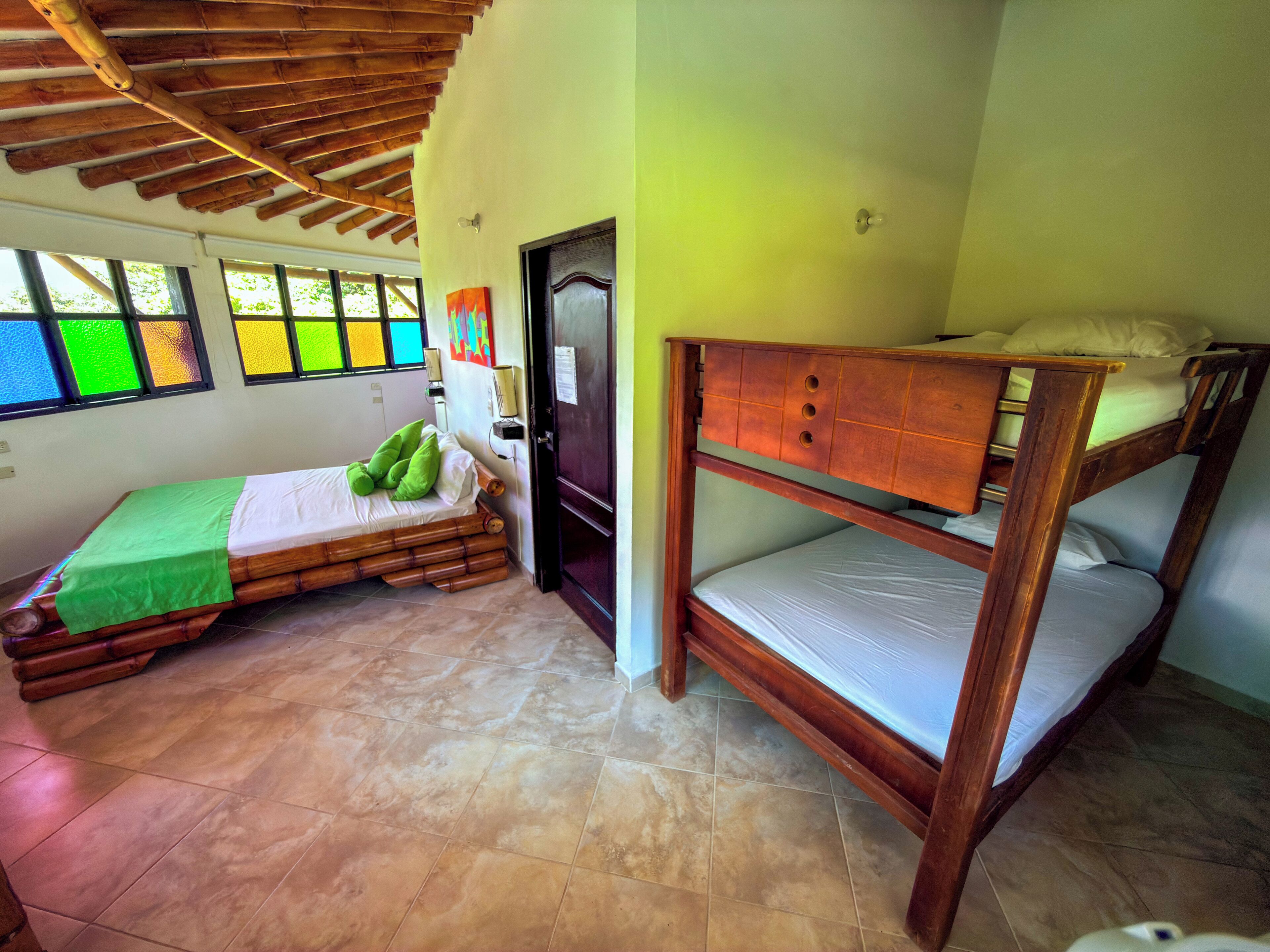 A room with a bunk bed, a single bed with a green bedspread, and a window with colorful panes.