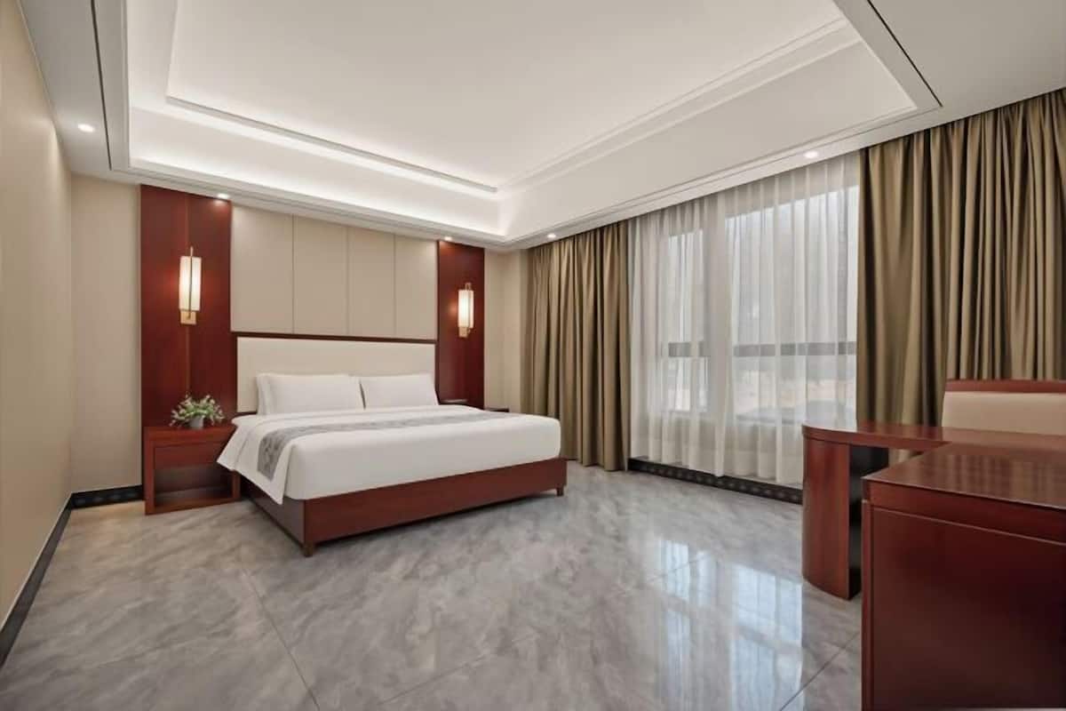 A modern hotel room with a large bed, a desk, and a view of the city through sheer curtains.