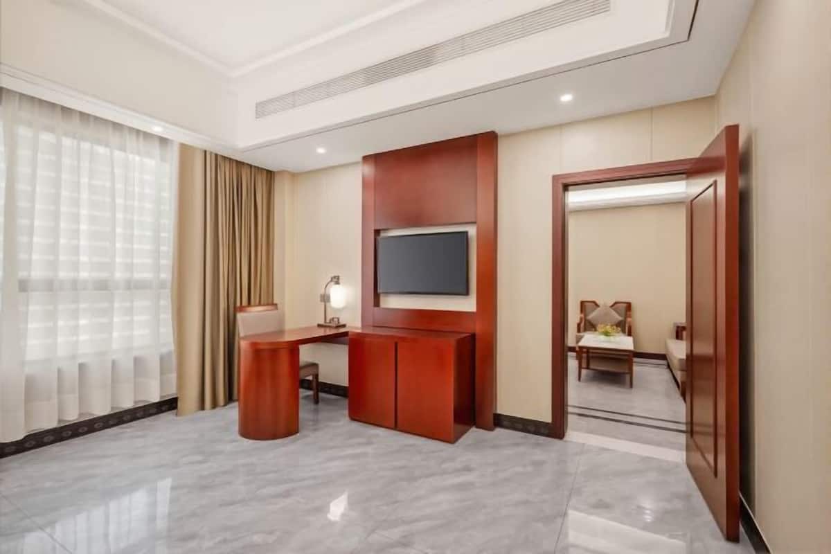 A hotel room with a flat-screen TV mounted on the wall, a desk with a lamp, a chair, and a dining area with a table and chairs.