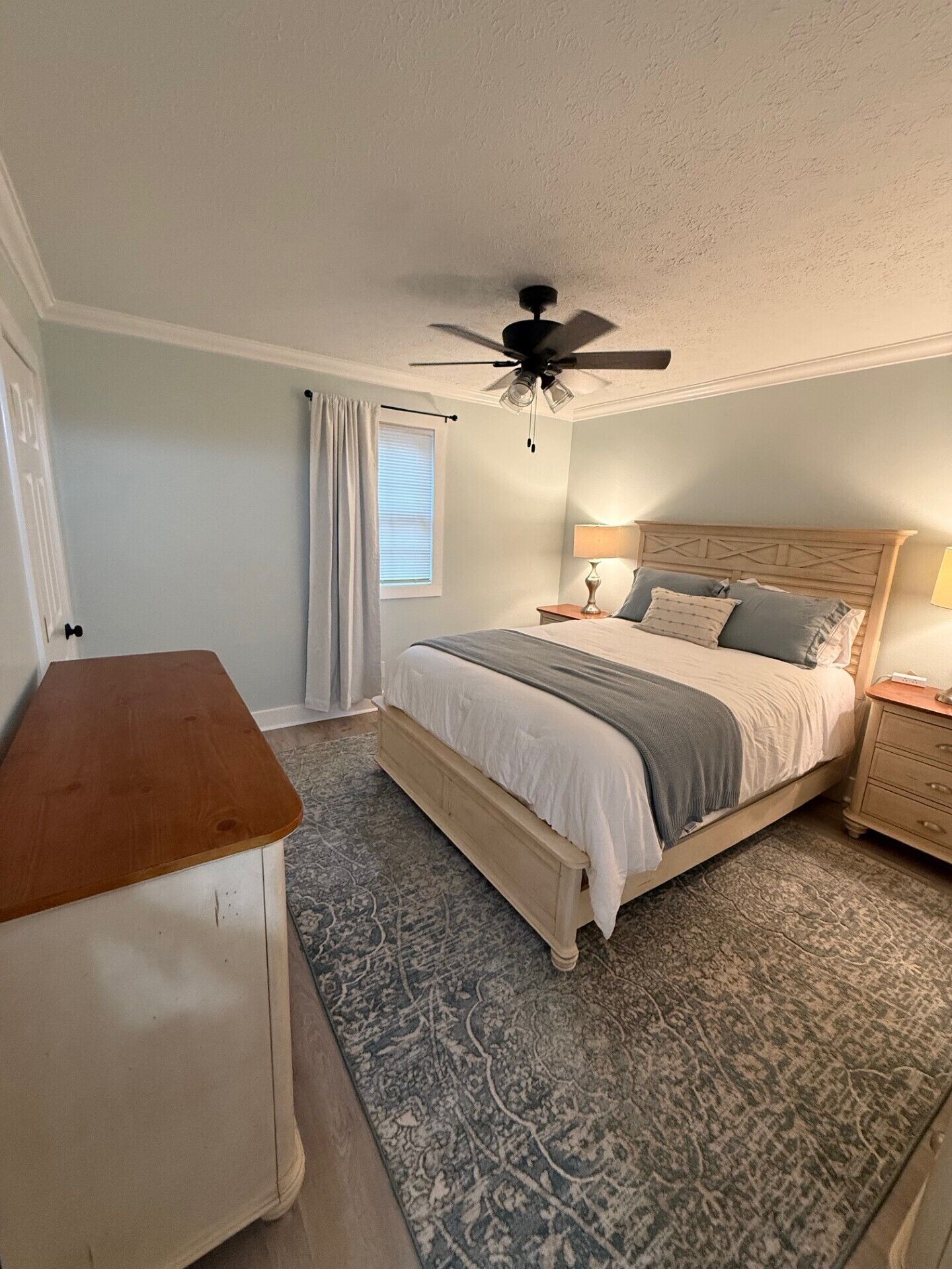 Une chambre à coucher comprenant un lit, une table de chevet, une commode en bois, un ventilateur de plafond et un tapis à motifs.
