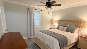Quarto com cama, mesa de cabeceira, cômoda de madeira, ventilador de teto e tapete estampado.