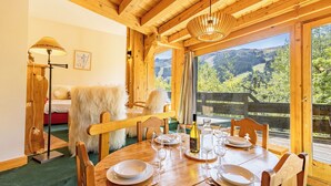 Une table à manger en bois, dressée avec des assiettes, des verres et une bouteille, et offrant une vue sur les montagnes à travers de grandes fenêtres.
