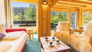 Une pièce chaleureuse avec un plafond en bois, un canapé rouge, une table en bois avec des tasses, et une vue sur les arbres et les montagnes.