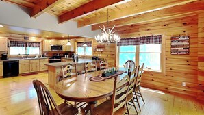 L’intérieur d’un chalet en bois comprend une table à manger entourée de chaises, un coin cuisine avec des placards en bois et un lustre.