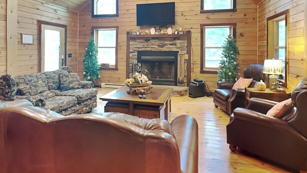 A log cabin interior with a fireplace, leather sofas, and Christmas trees. - Log Heaven by Taylor Made Deep Creek (Oakland)