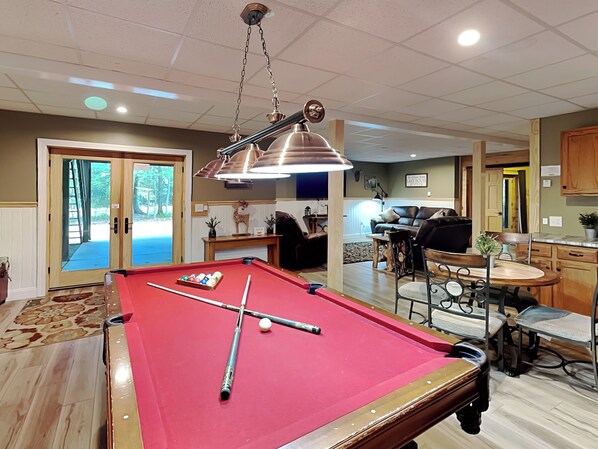 Une table de billard avec des billes et des queues, un coin repas avec une table et des chaises, un espace cuisine avec des placards, et un salon avec un canapé et une table basse.