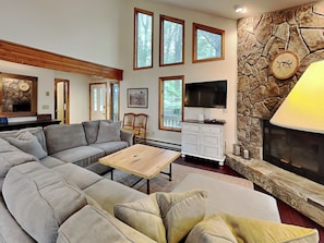 A modern living room with a sectional sofa, a flat-screen TV, a stone fireplace, and large windows. - Somewhere by Taylor Made Deep Creek (Oakland)
