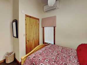 A bedroom with a wooden door, a window with blinds, a bed with a floral bedspread, and an air conditioning unit on the wall.