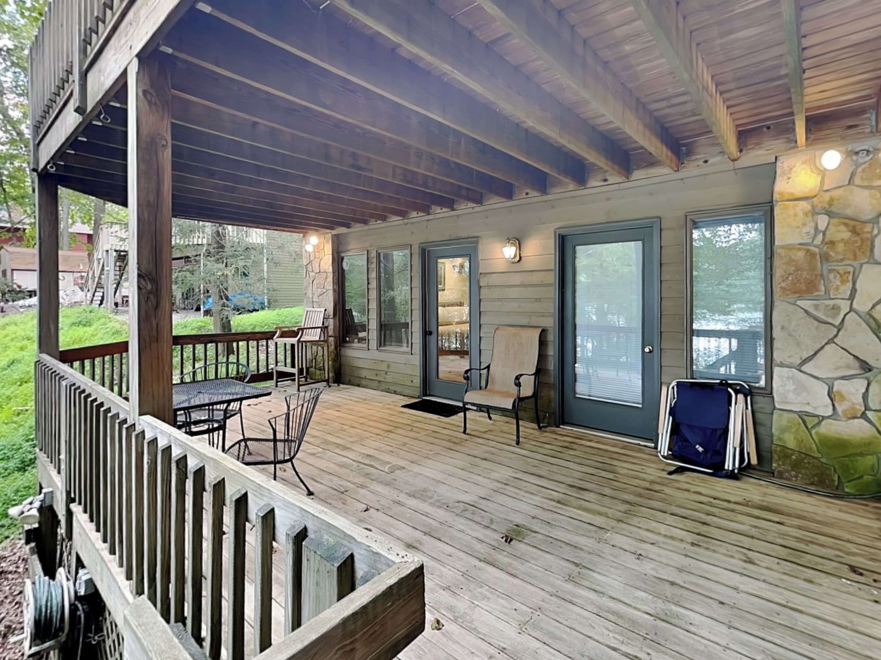 A covered outdoor deck with a wooden floor, a dining table with chairs, and a stone wall.