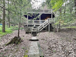 A wooden deck with a staircase leading to a covered area, surrounded by trees and a gravel path. - Somewhere by Taylor Made Deep Creek (Oakland)