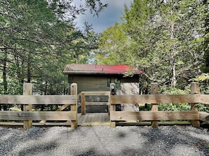 A rustic cabin with a red roof, surrounded by a wooden fence and dense forest.