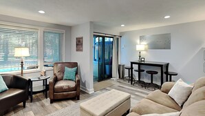 A modern living room with a leather armchair, a beige sofa, a small table with a lamp, and a dining area with stools. - Arrowhead Village 13 by Taylor Made Deep Creek (Oakland)