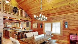 A spacious wooden cabin with a large dining table, multiple chairs, a kitchen area, and a balcony with a dog. - Crow's Point Lakehouse by Taylor Made Deep Creek (Swanton)