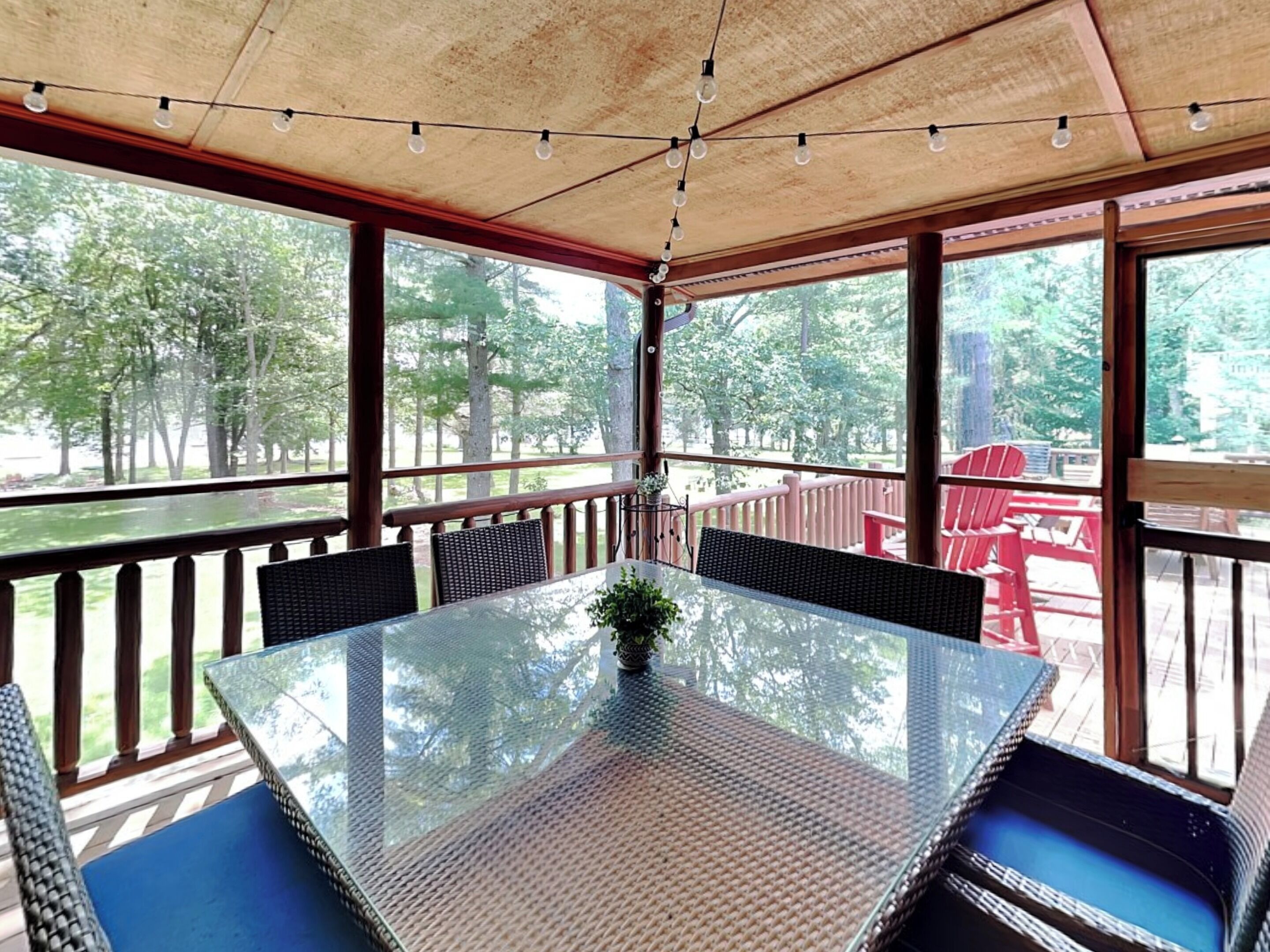 A covered outdoor patio with a glass table and wicker chairs, overlooking a deck with red chairs and a view of trees.