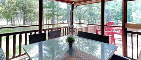 A covered outdoor patio with a glass table and wicker chairs, overlooking a deck with red chairs and a view of trees.